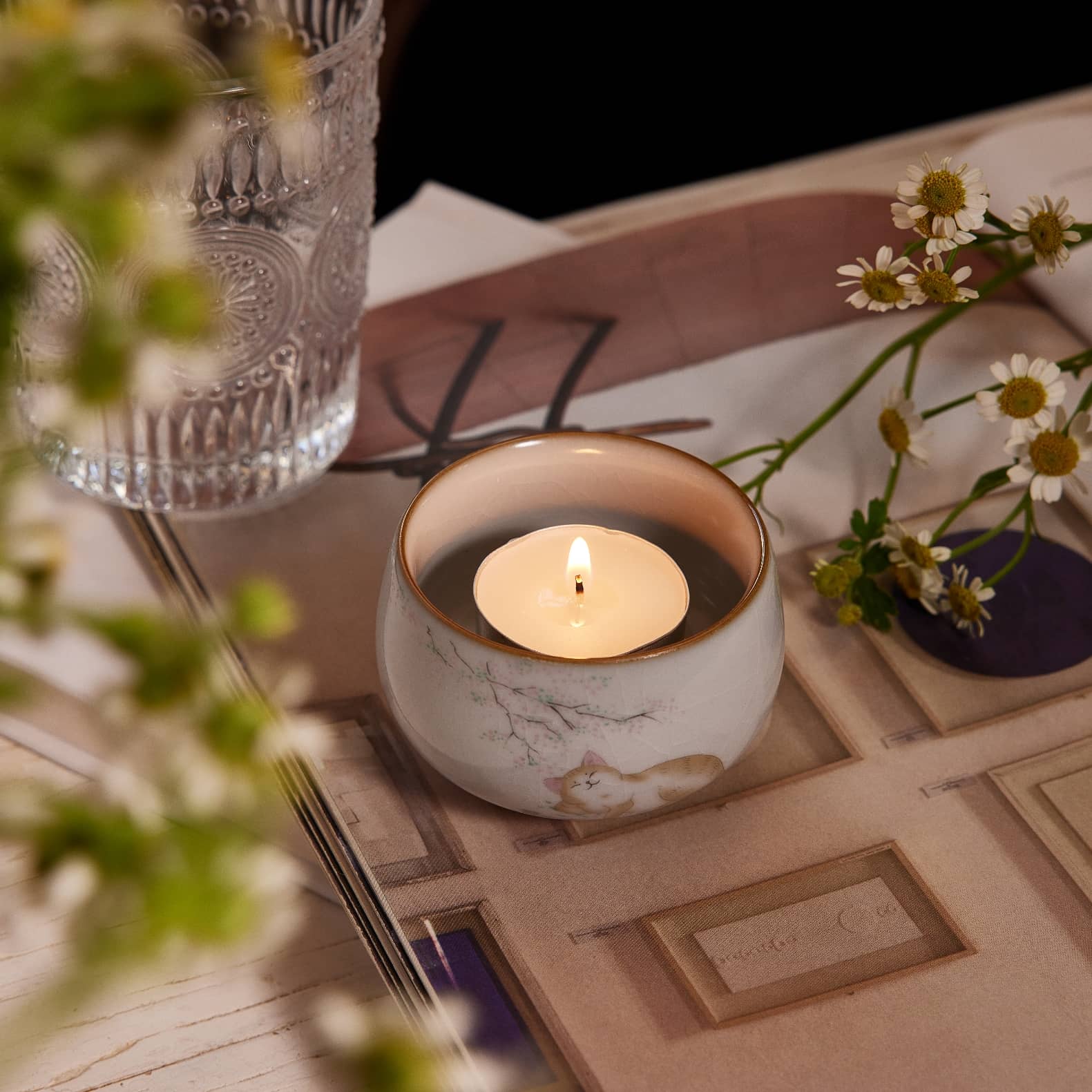 A lighting tealight inside a tealight candle holder with an adorable orange cat lay down under a cherry blossom, looks very chill and enjoy the vibes.